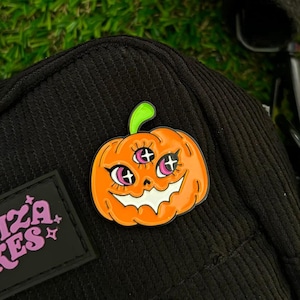 May include: Orange pumpkin with a spooky face and four eyes. The pumpkin has a pink and white smile and pink eyelids. The pumpkin is a pin on a black background.
