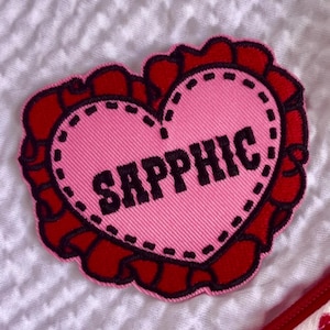 Könnte beinhalten: Ein herzförmiger Aufnäher in Rosa mit dem Wort „SAPPHIC“ in schwarzer Schrift. Das Herz ist mit einer schwarzen gepunkteten Linie und einem roten, gewellten Rand umrandet. Zum Aufnähen auf Kleidung.