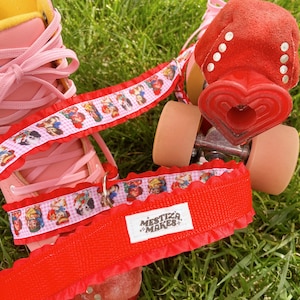 May include: Pink roller skates with red wheels and a red heart-shaped toe stop. The skates have a white and pink gingham ribbon with cartoon characters printed on it. The ribbon is tied around the skates and has a red ribbon with the text "Mestiza Makes" printed on it.