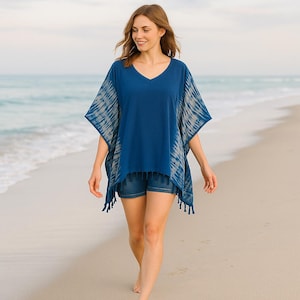 Può includere: Un poncho blu con scollo a V con maniche tie-dye e finiture con frange. La modella indossa pantaloncini di jeans e cammina su una spiaggia sabbiosa. L'oceano è visibile sullo sfondo.