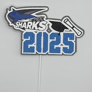 May include: Blue and gray graduation cake topper with a shark, graduation cap, and diploma. The topper says "MDC Sharks 2025".