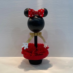 Pode incluir: Figura de Minnie Mouse preta e vermelha com um la&ccedil;o vermelho e bolinhas brancas. A figura est&aacute; usando um vestido vermelho com luvas brancas e uma fita dourada. A figura est&aacute; em p&eacute; sobre uma base vermelha com um vaso preto.