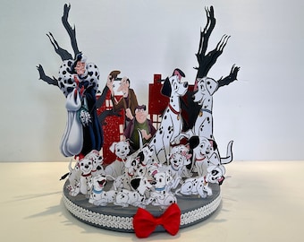 Può includere: Una scena di papercraft 3D con personaggi del film Disney 101 Dalmatiani. Cruella De Vil, Roger, Anita e i cuccioli dalmata sono rappresentati in uno schema di colori bianco e nero. La scena è ambientata sullo sfondo di uno skyline cittadino e alberi. Un fiocco rosso è posto al centro della base.