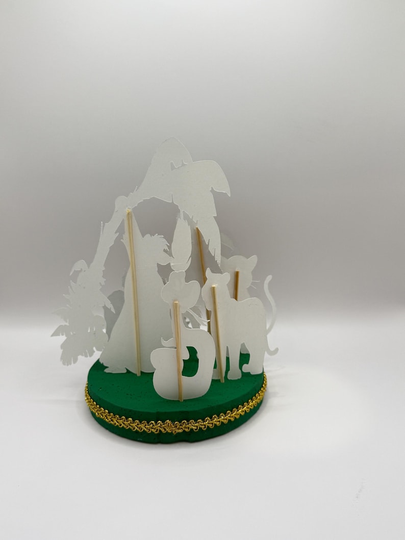 Peut inclure: Un ensemble de marionnettes d'ombres en papier blanc avec un palmier, un chat, un chien et la lettre D. Les marionnettes sont mont&eacute;es sur des b&acirc;tonnets en bois et sont pos&eacute;es sur une base circulaire verte avec une bordure dor&eacute;e.