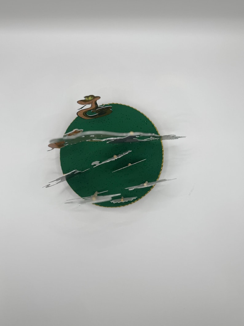 Peut inclure: Une carte pop-up 3D ronde en papier de couleur verte avec une bordure dor&eacute;e. La carte repr&eacute;sente un serpent avec une t&ecirc;te verte et une couronne dor&eacute;e. Le serpent est sur une base verte avec des nuages blancs.