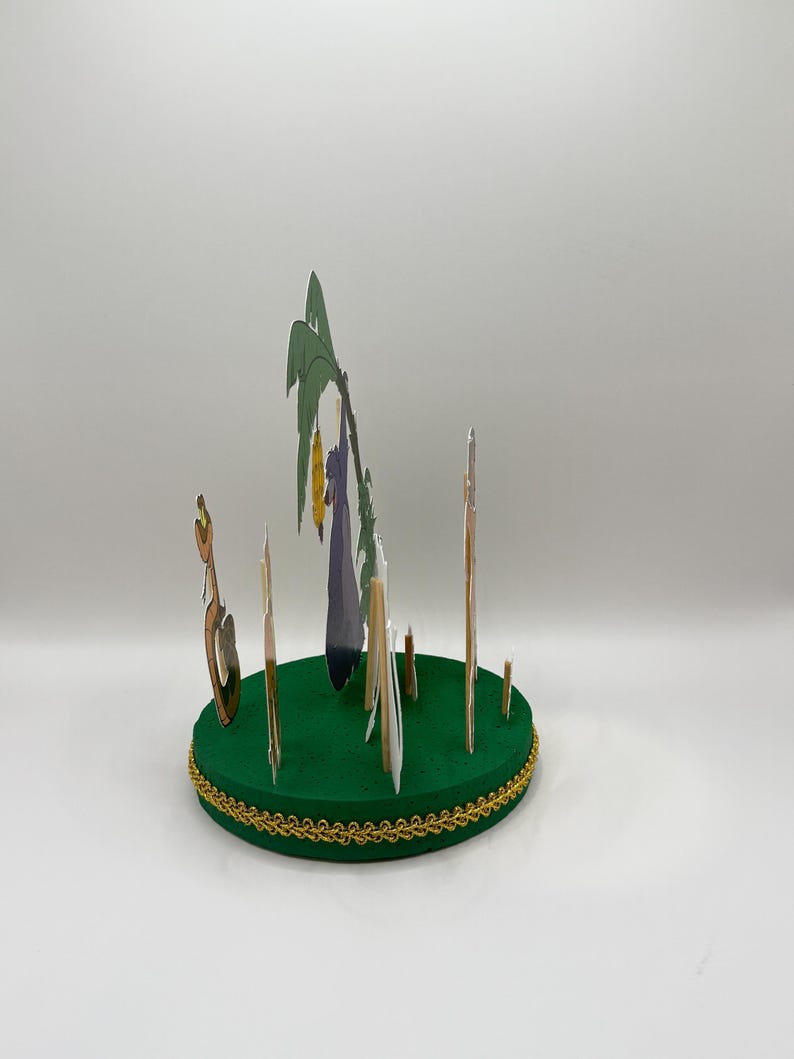 Peut inclure: Une sc&egrave;ne en papier d&eacute;coup&eacute; verte et dor&eacute;e avec un palmier, une figurine violette et plusieurs b&acirc;tonnets bruns. La sc&egrave;ne est sur une base circulaire verte.