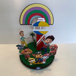 Puede incluir: Un adorno para pastel con tem&aacute;tica de la Patrulla Canina con un arco&iacute;ris, un faro y personajes del programa, incluidos Ryder, Chase, Skye y Marshall.