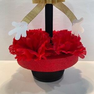 Pode incluir: Centro de mesa Mickey Mouse preto e vermelho com fita dourada e m&atilde;os de papel branco. O centro de mesa &eacute; decorado com cravos vermelhos.