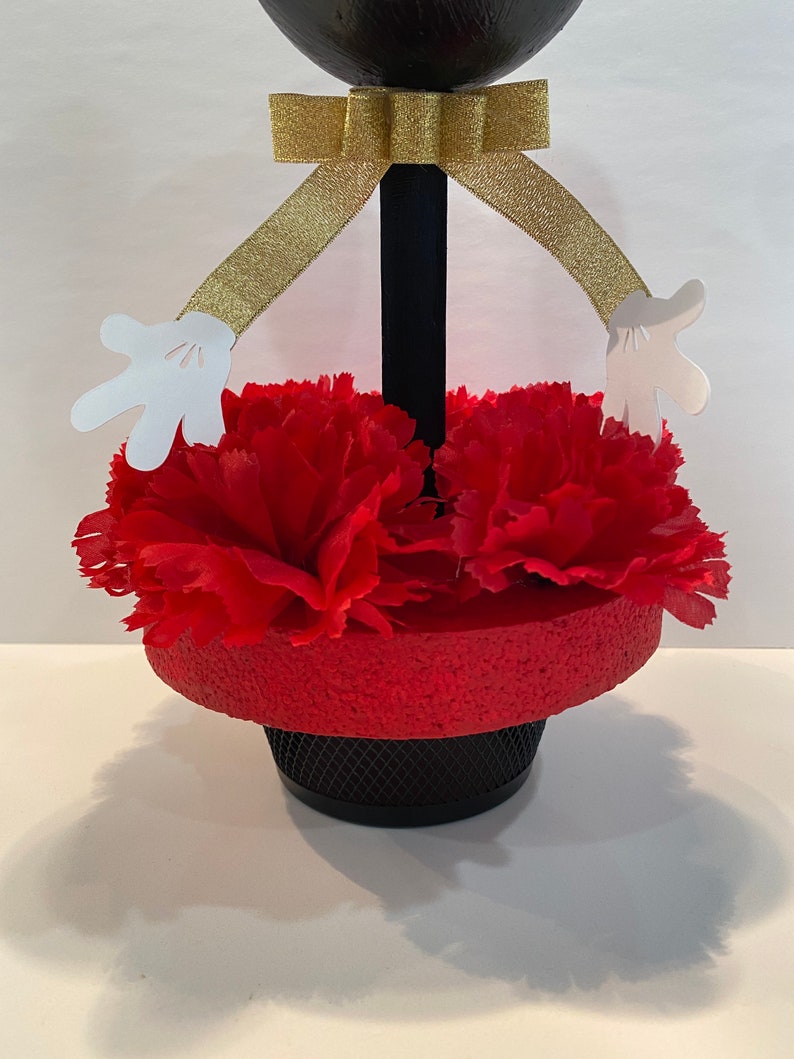 Pode incluir: Centro de mesa Mickey Mouse preto e vermelho com fita dourada e m&atilde;os de papel branco. O centro de mesa &eacute; decorado com cravos vermelhos.
