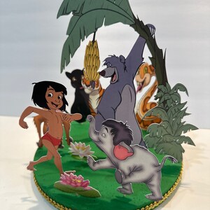 Peut inclure: Une sc&egrave;ne en papier 3D repr&eacute;sentant des personnages du Livre de la Jungle. Mowgli, Baloo l'ours, Shere Khan le tigre, Kaa le serpent et un &eacute;l&eacute;phant se tiennent sur une base verte avec un palmier et des fleurs roses.