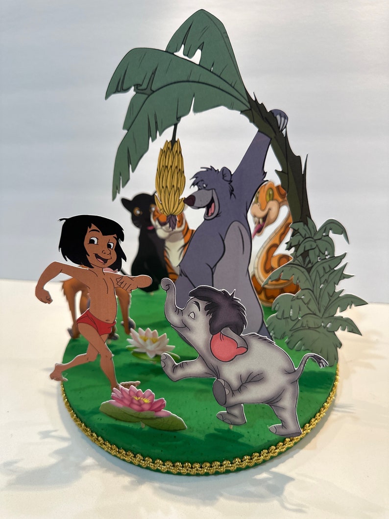 Peut inclure: Une sc&egrave;ne en papier 3D repr&eacute;sentant des personnages du Livre de la Jungle. Mowgli, Baloo l'ours, Shere Khan le tigre, Kaa le serpent et un &eacute;l&eacute;phant se tiennent sur une base verte avec un palmier et des fleurs roses.