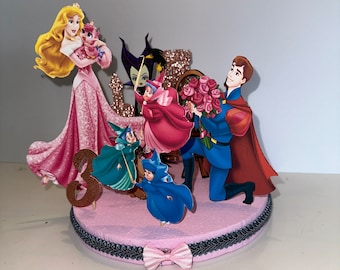 Könnte beinhalten: Ein rosa Tortenaufsatz mit Figuren aus dem Disney-Film Dornröschen. Der Aufsatz enthält Dornröschen, Prinz Philipp, Maleficent, die drei Feen und eine rosa Zahl 3.