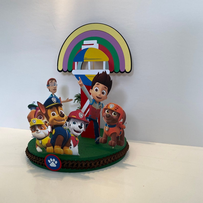 Puede incluir: Un adorno para pastel con tem&aacute;tica de la Patrulla Canina con un arco&iacute;ris, una torre de vigilancia y personajes como Chase, Marshall, Skye y Ryder. El adorno est&aacute; hecho de cart&oacute;n y presenta detalles coloridos.