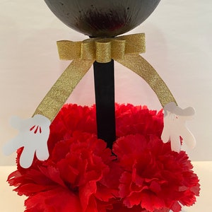 Pode incluir: Centro de mesa com cabe&ccedil;a de Mickey Mouse em preto e branco com um la&ccedil;o dourado e cravos vermelhos. A cabe&ccedil;a do Mickey Mouse &eacute; feita de uma bola preta redonda com luvas brancas presas aos lados.