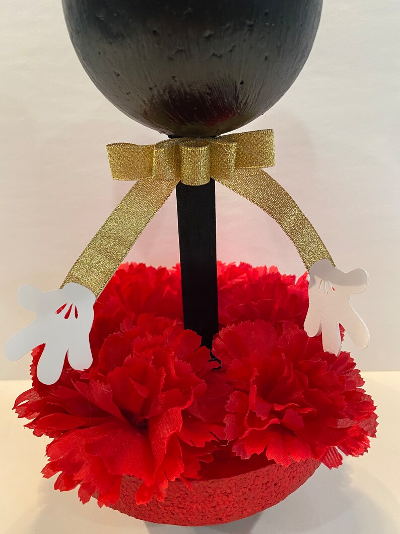 Pode incluir: Centro de mesa com cabe&ccedil;a de Mickey Mouse em preto e branco com um la&ccedil;o dourado e cravos vermelhos. A cabe&ccedil;a do Mickey Mouse &eacute; feita de uma bola preta redonda com luvas brancas presas aos lados.