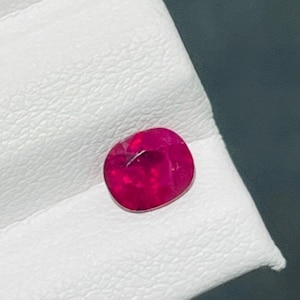 Può includere: Una gemma rosso vivo e profondo con taglio a cuscino, caratterizzata da molteplici sfaccettature che riflettono la luce sulla sua superficie. La pietra presenta una ricca tonalità cremisi satura ed è posizionata su una base bianco sporco.