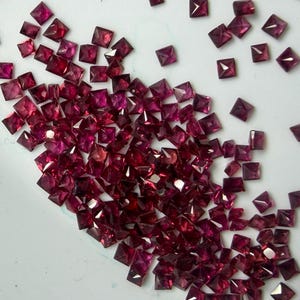 Puede incluir: Una colección de pequeñas gemas cuadradas, de color rojo intenso, dispersas sobre una superficie blanca. Las gemas tienen una apariencia facetada, captando la luz y mostrando una gama de tonos que van desde el rubí hasta un tono más oscuro, casi púrpura. Las gemas son de varios tamaños.