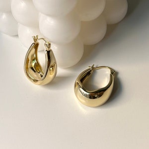May include: Two gold-colored, teardrop-shaped hoop earrings. The earrings have a smooth, polished surface and a rounded, chunky design. They are displayed on a white surface, with a white, spherical decorative object in the background.