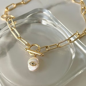 May include: Gold chain necklace with a toggle clasp and a white pearl charm with a gold evil eye design.