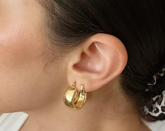 May include: A pair of gold-colored hoop earrings. The earrings are thick and rounded, with a polished, reflective surface. The hoops are secured with a hinged clasp. The earrings are shown on an ear.