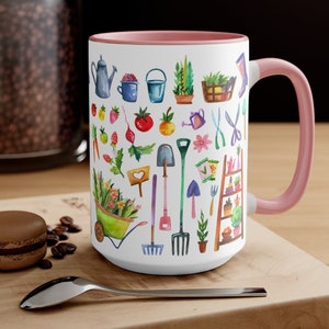 May include: White ceramic mug with a pink handle and interior. The mug features a watercolor design of gardening tools, plants, and vegetables. A spoon and a macaron are in the foreground.