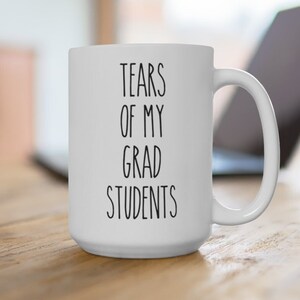 May include: White ceramic coffee mug with black text that reads "Tears of my grad students".