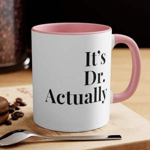 May include: White ceramic coffee mug with a pink rim and handle. The mug has black text that reads "It's Dr. Actually".