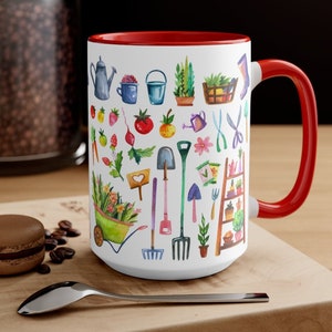 May include: White ceramic mug with a red interior and handle, featuring a watercolor design of gardening tools, plants, and produce. The mug includes a watering can, wheelbarrow, and various gardening implements. The mug is sitting on a wooden surface.