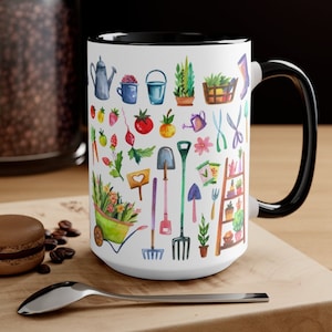 May include: White ceramic mug with a black rim and handle. The mug features a colorful pattern of gardening tools, plants, and vegetables.