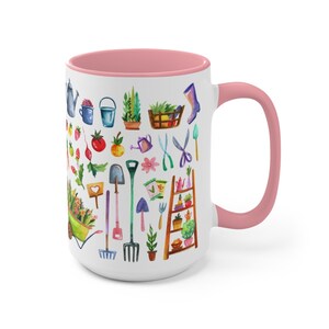 May include: White ceramic mug with a pink interior and handle. The mug is decorated with watercolor illustrations of gardening tools, plants, and vegetables. The design includes a wheelbarrow, watering can, and a small wooden ladder with potted plants.