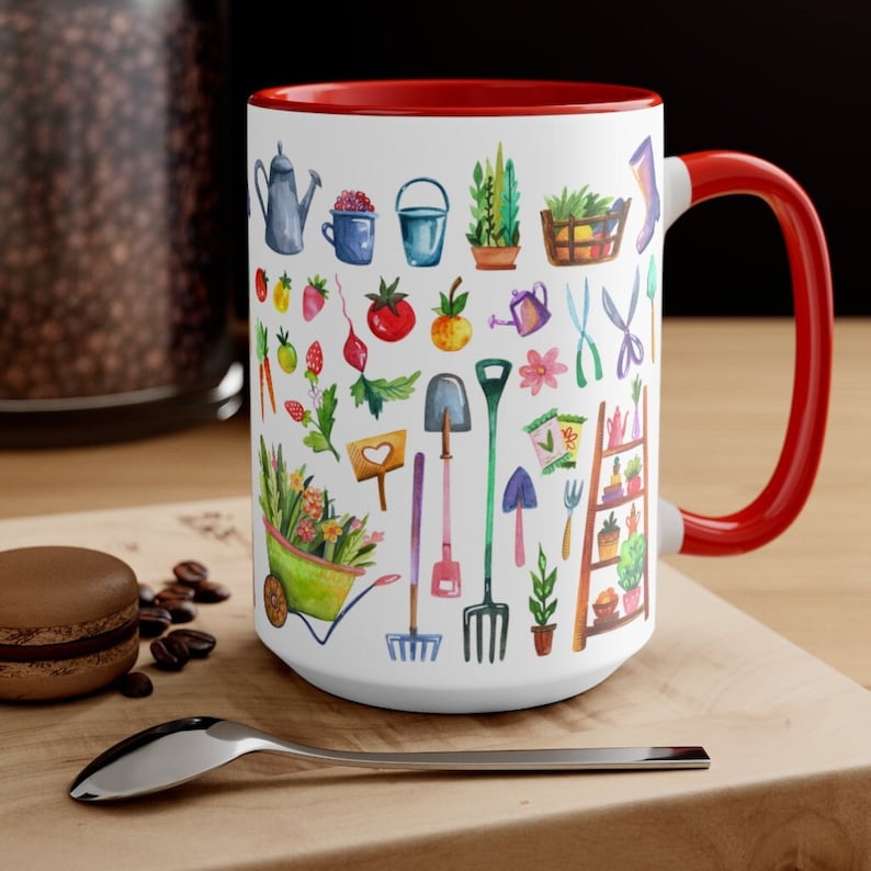 May include: White ceramic mug with a red interior and handle, featuring a watercolor design of gardening tools, plants, and produce. The mug includes a watering can, wheelbarrow, and various gardening implements. The mug is sitting on a wooden surface.