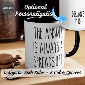 May include: White ceramic mug with a black handle. The mug has black text that reads "The answer is always a spreadsheet". The mug is on a wooden surface with coffee beans and a macaron.