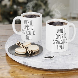 May include: Two white coffee mugs with black text that reads "When it comes to spreadsheets I excel". The mugs are on a white marble surface with a silver plate and macarons.