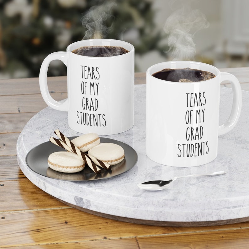 May include: Two white coffee mugs with black text that reads "Tears of my grad students". The mugs are filled with coffee and steam is rising from the mugs. The mugs are on a white marble surface with a silver plate and macarons.