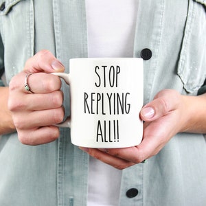 May include: A white ceramic mug with the text "STOP REPLYING ALL!!" in black, held by a person. The mug is the focal point, with the text prominently displayed. The person is wearing a light green jacket.
