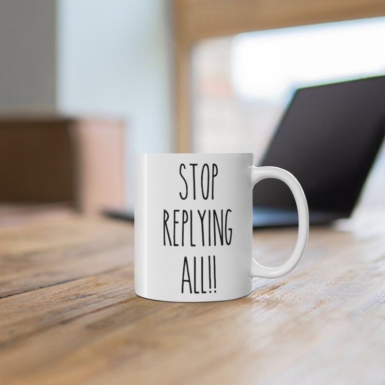 May include: White ceramic mug with the text "STOP REPLYING ALL!!" in black, set on a wooden surface. A laptop is visible in the background, slightly out of focus.