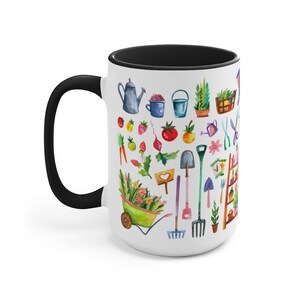 May include: White ceramic mug with a black rim and handle. The mug features a watercolor illustration of gardening tools, plants, and flowers. The design includes a watering can, a wheelbarrow, a shovel, a rake, a trowel, a pair of gardening shears, a tomato, a strawberry, a carrot, a flower, and a potted plant.