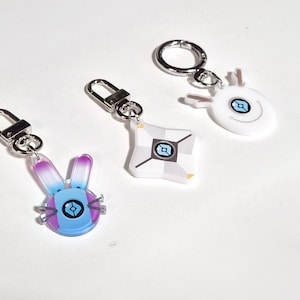 Peut inclure: Trois porte-clés avec des attaches argentées. L'un présente un design de lapin bleu, violet et blanc. Un autre a une forme géométrique blanche et grise. Le troisième porte-clés a un design ovale blanc.