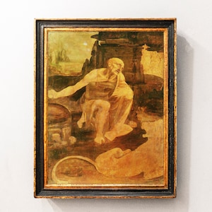 May include: A framed painting depicting a bearded man in a robe sitting with a lion. The painting is set in a landscape with a rocky background and a golden hue.