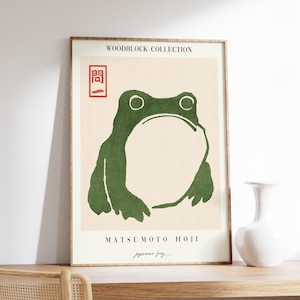 Op de afbeelding: Een ingelijste print van een groene kikker met een rode zegel waarop in het Japans "関" staat. De print is getiteld "MATSUMOTO HOJI" en heeft de tekst "Japanese Frog" eronder.