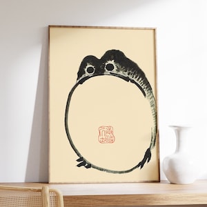 Könnte beinhalten: Ein Schwarzweiß-Tuschbild eines Frosches in einem Kreis, mit einem roten Siegel in der Mitte. Das Siegel enthält japanische Schriftzeichen. Das Bild ist auf cremefarbenem Hintergrund und in einem hellen Holzrahmen gerahmt.