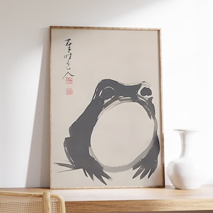 Könnte beinhalten: Eine Schwarzweiß-Tuschzeichnung eines Frosches in einem einfachen, minimalistischen Stil. Die Zeichnung ist gerahmt und hängt an einer weißen Wand. Die japanischen Schriftzeichen "蛙" (Frosch) und "雨" (Regen) sind in der oberen linken Ecke der Zeichnung geschrieben.
