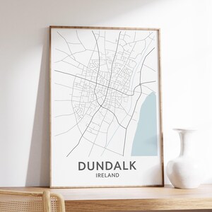 Może przedstawiać: Oprawiony wydruk z minimalistyczną mapą Dundalk, Irlandia, z jasnoniebieskim obszarem przybrzeżnym. Mapa jest w czarnych liniach na białym tle, z nazwą miasta i kraju wydrukowanymi poniżej ciemnoszarą czcionką. Rama jest w kolorze jasnobrązowym.