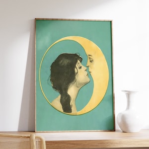 Op de afbeelding: Een ingelijste print van een vrouw die een wassende maan kust. De vrouw heeft donker haar en draagt een witte jurk. De maan is geel en heeft een menselijk gezicht. De achtergrond is turquoise.