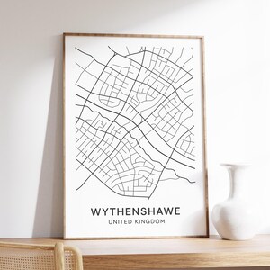 Peut inclure: Une impression encadrée en noir et blanc de la carte de Wythenshawe, Royaume-Uni. La carte présente un plan détaillé des rues. L'impression est dans un cadre en bois et repose sur une surface en bois. Un vase blanc est à droite de l'impression.