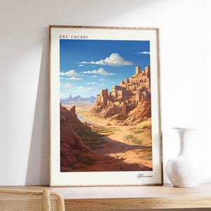 May include: Framed art print featuring a desert landscape with a village built into a hillside. The artwork has a warm color palette with a blue sky and the text "ERG CHEBBI" at the top. A white vase sits on a wooden surface.