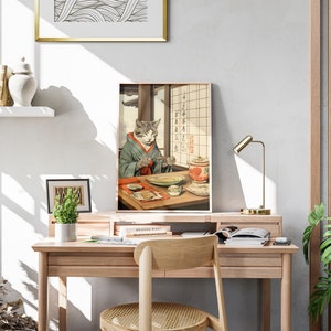 Puede incluir: Una impresi&oacute;n enmarcada de un gato con un kimono y sosteniendo palillos, sentado en una mesa con un taz&oacute;n de comida y una tetera. La impresi&oacute;n est&aacute; en un escritorio de madera con una l&aacute;mpara, libros y plantas.