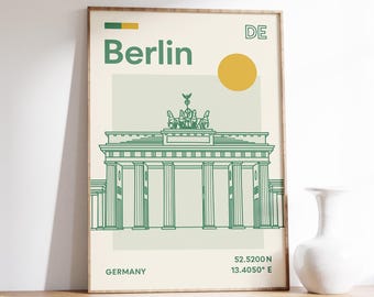 May include: A framed art print featuring a green and cream illustration of the Brandenburg Gate in Berlin. The word "Berlin" is above the gate, with the country code "DE" and coordinates. A yellow sun graphic is also included.