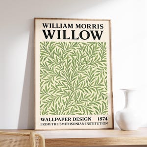 Op de afbeelding: Een ingelijste print met het William Morris "Willow" behangontwerp uit 1874. Het kunstwerk toont een herhalend patroon van groene wilgenbladeren op een crèmekleurige achtergrond. De print wordt weergegeven op een houten oppervlak, met een witte vaas en een stoel zichtbaar.
