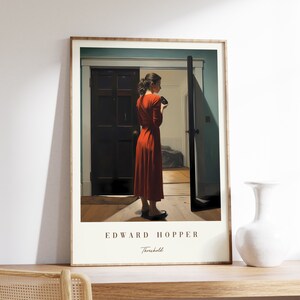Pode incluir: Uma impressão emoldurada de uma pintura de Edward Hopper intitulada "Threshold". A pintura retrata uma mulher com um vestido vermelho em pé em uma porta, olhando para algo em sua mão.
